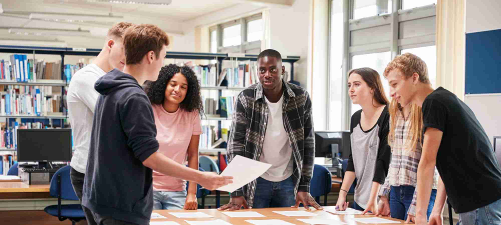 Six students gather around a library table, reviewing papers together in a bright, collaborative setting.